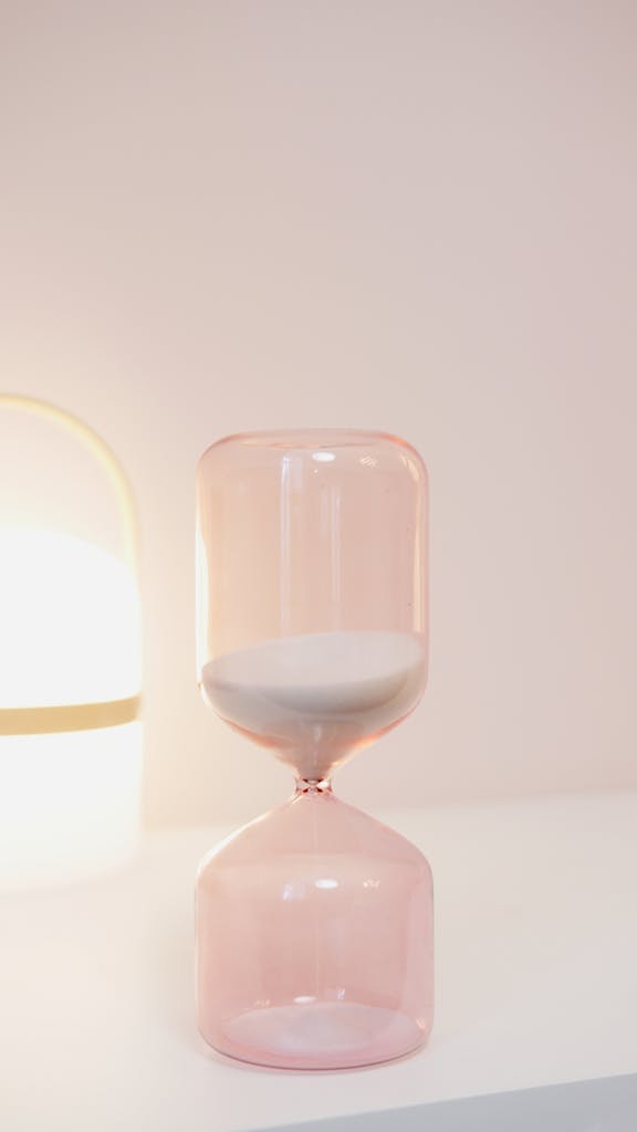 Elegant pink hourglass on a white shelf with soft lighting, ideal for design and time concept visuals.